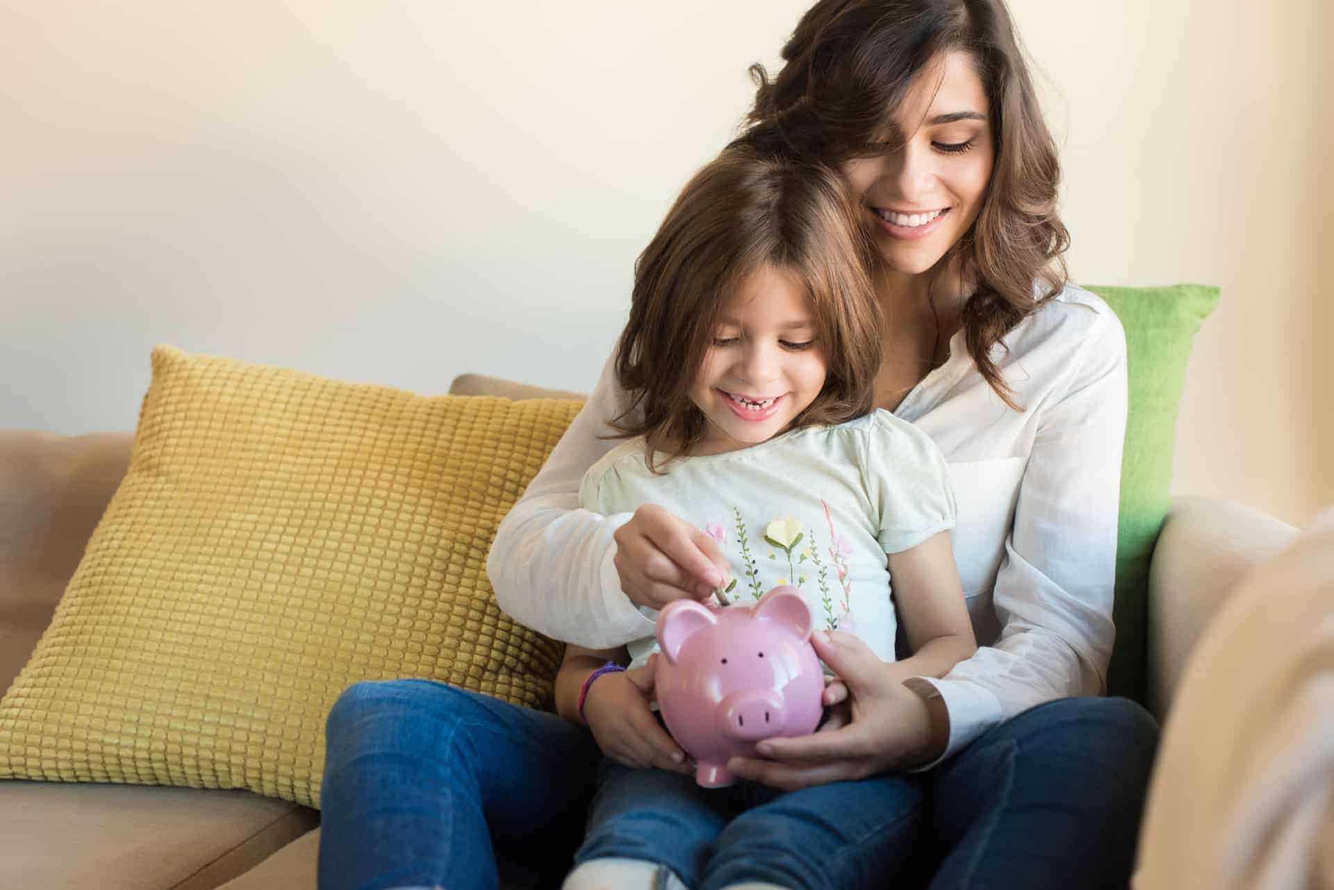 Woman holding a child who is holding a piggy bank. They are putting money into the piggy bank.