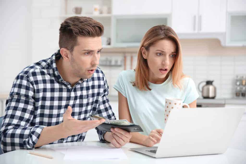 Young man holding wallet next to a woman holding a coffee cup while both are upset while looking at a computer screen