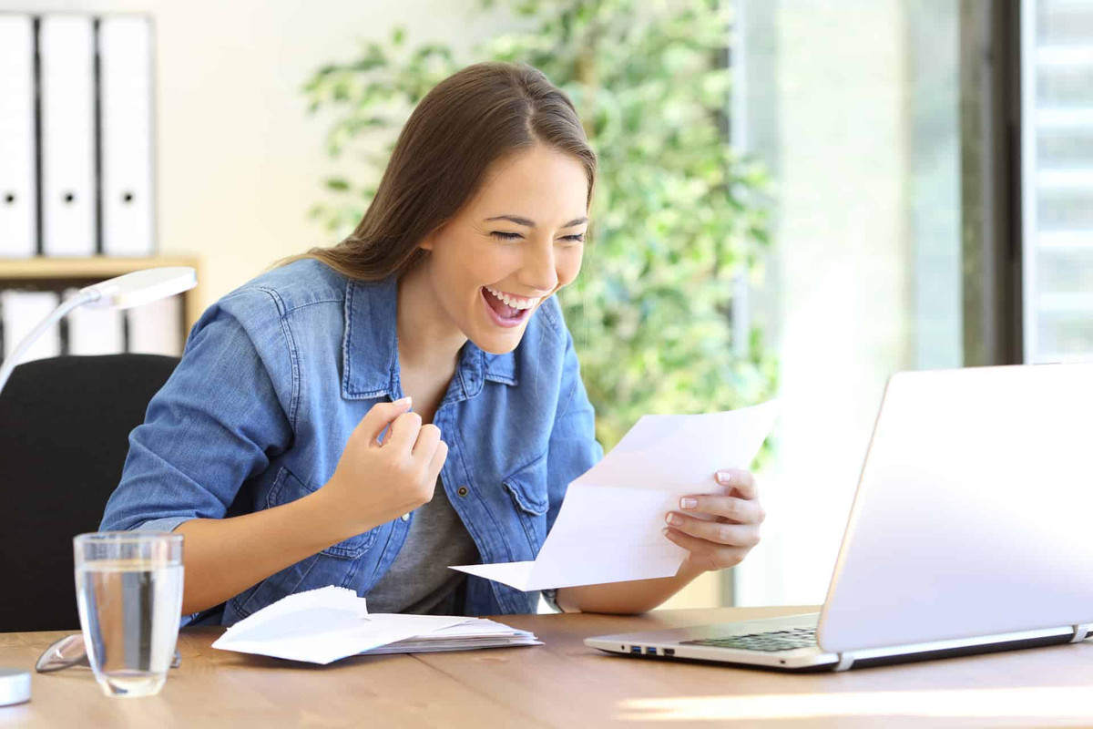 Excited woman holding a piece of paper and looking at her computer.