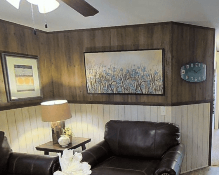Image of a living room with half wooden walls and half beadboard. There is one full recliner pictured in the scene, and some wall art. Next to the recliner is a small table with a lamp and a few decorative pieces on top.