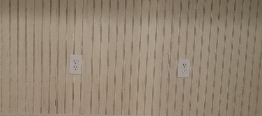 Image of beadboard on wall with two outlets, there are signs of wear and scratches all over the beadboard wall.